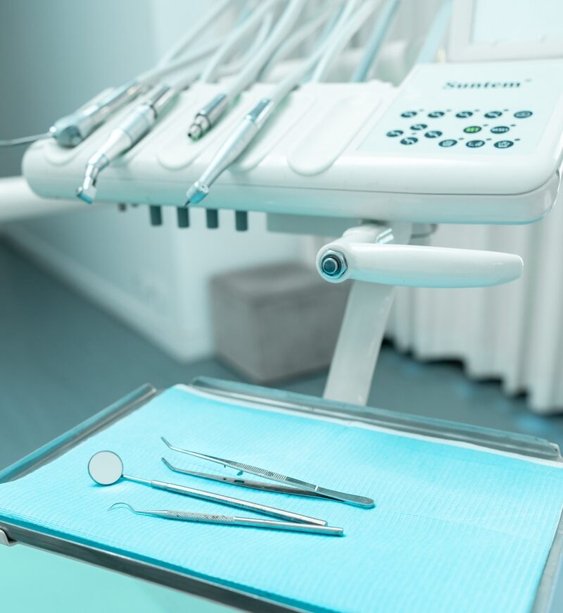 A close-up of a dental workstation with various stainless steel dental tools neatly arranged on a blue sterile pad, alongside a modern dental unit with handpieces and control buttons. - Total Health Environment