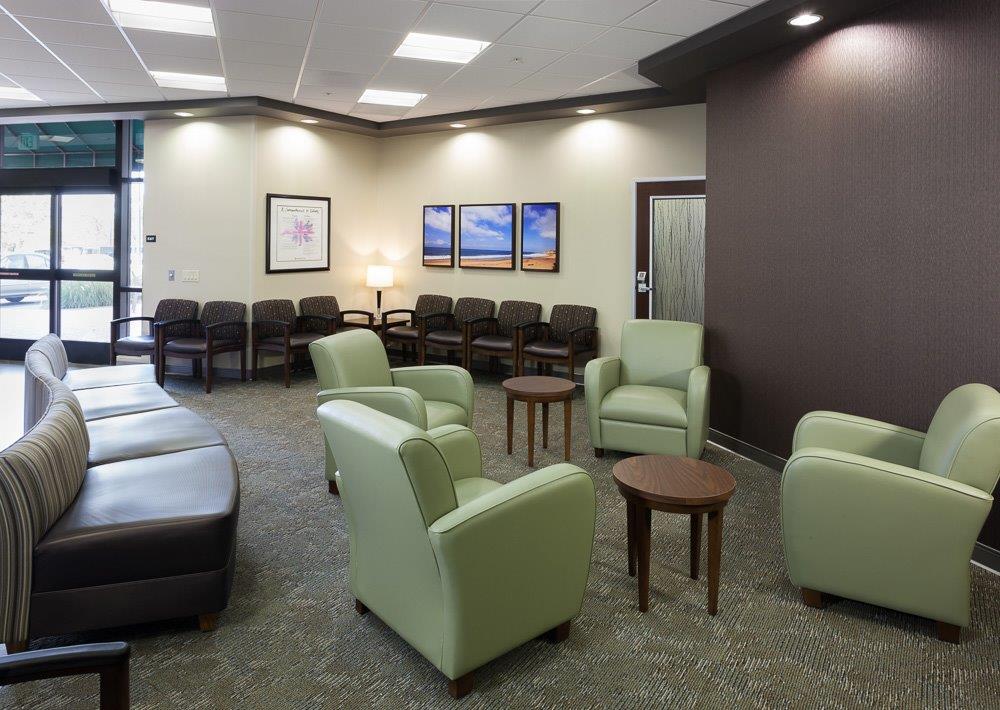 A modern medical waiting area with green and brown seating, round tables, soft lighting, and framed artwork on the walls, creating a calm and inviting atmosphere. - Total Health Environment
