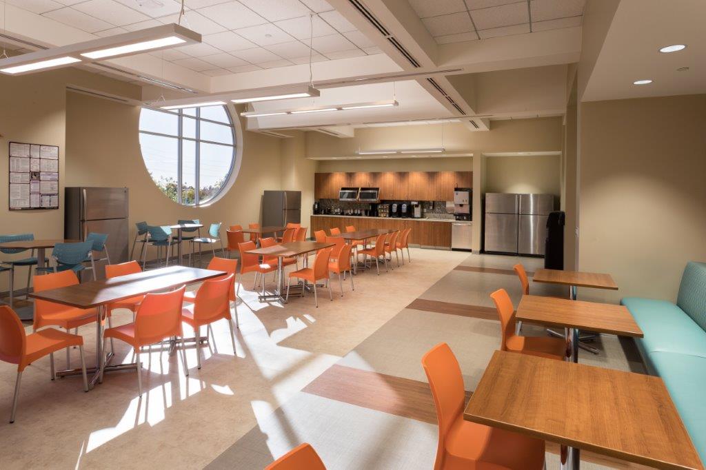 A modern and well-lit employee break room with bright orange chairs, wooden tables, stainless steel refrigerators, microwaves, and a coffee station, featuring a large circular window allowing natural light. - Total Health Environment