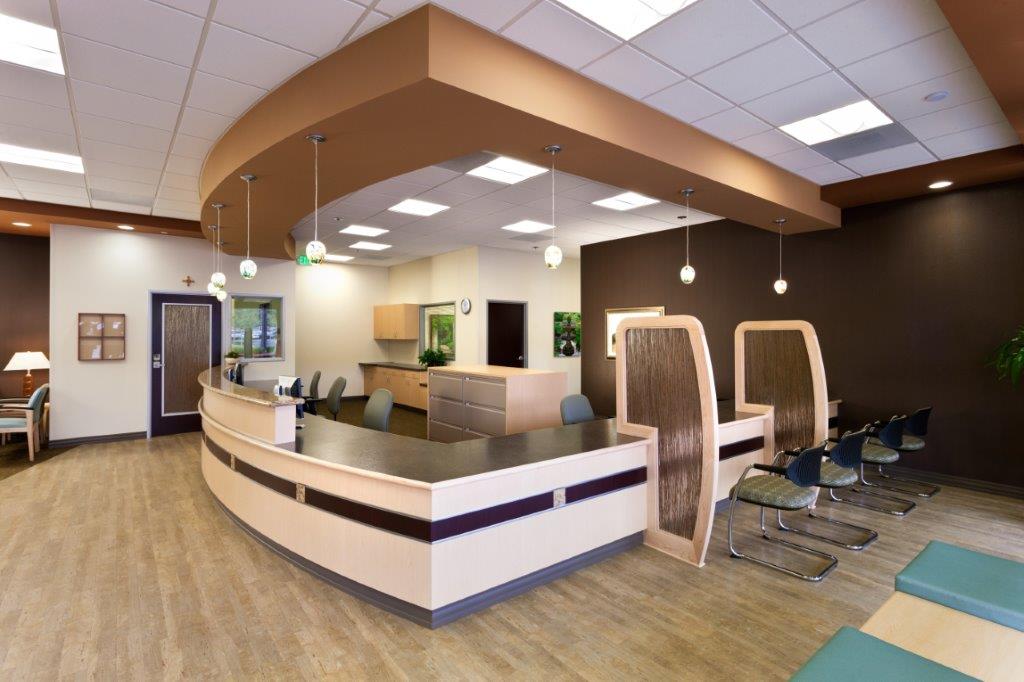 A spacious and well-lit healthcare reception area features a curved front desk, seating for patients, pendant lighting, and a warm color palette with wood and neutral tones. - Total Health Environment