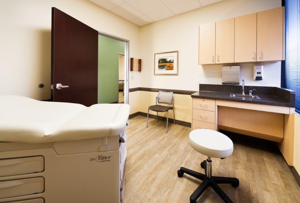 A clean and organized medical exam room features a padded examination table, a stool, a chair for visitors, wooden cabinets, a sink, and a framed landscape painting on the wall. - Total Health Environment