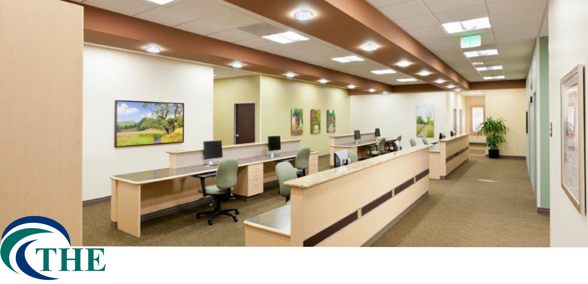 A spacious and well-lit medical office reception area featuring a long front desk, multiple workstations, framed landscape artwork, and a logo in the bottom left corner reading "THE" for Total Health Environment. - Total Health Environment