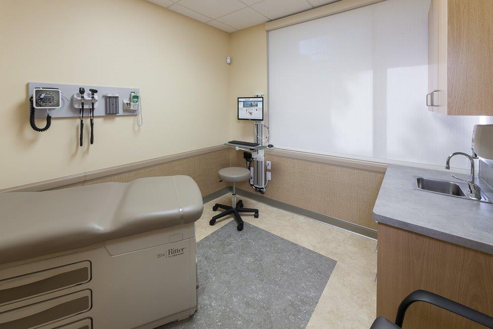  A clean and well-organized medical examination room features an exam table, a stool, a computer workstation, and medical equipment mounted on the wall, creating a professional and functional healthcare setting. - Total Health Environment